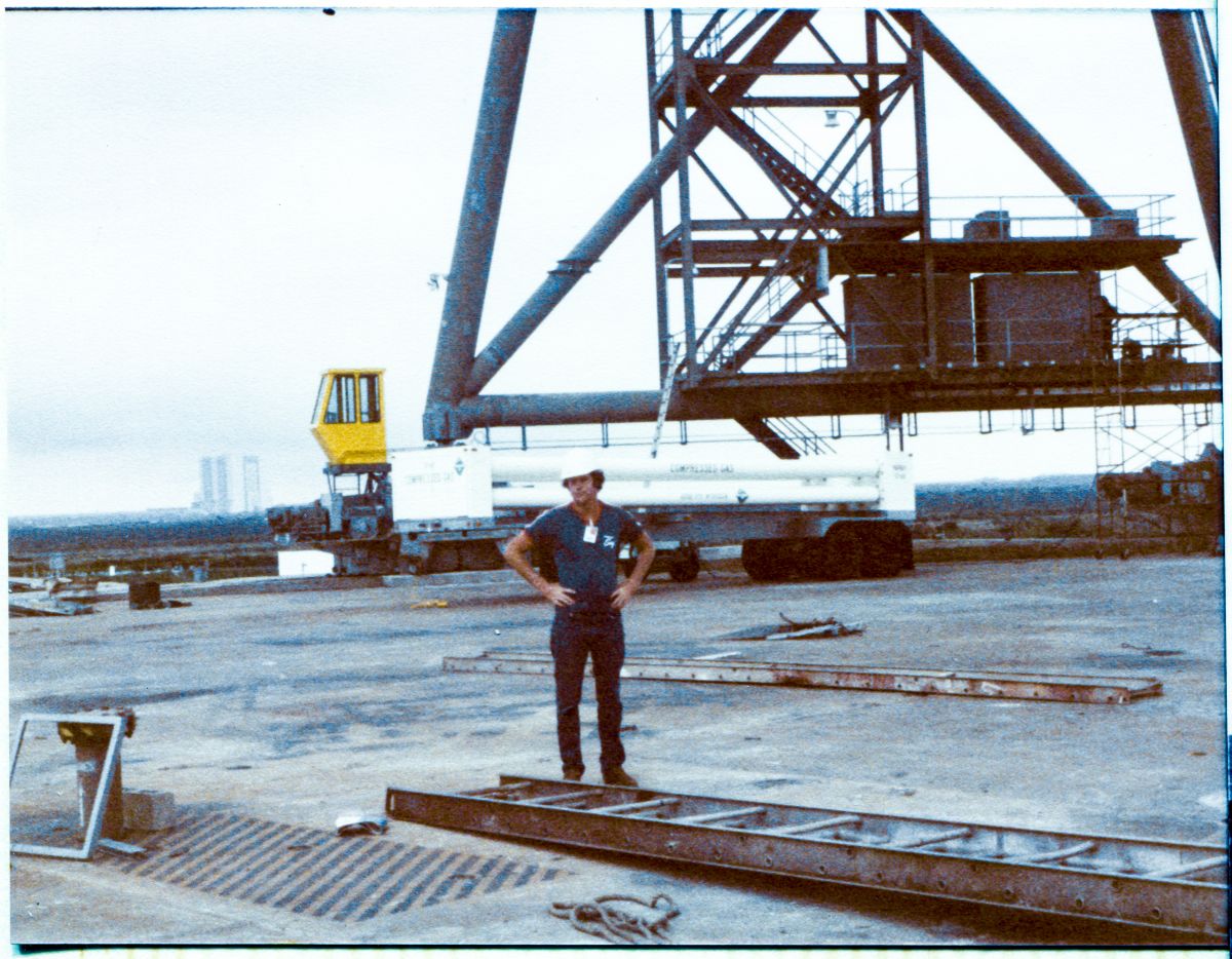 Image 018. James MacLaren stands on the Pad Deck beneath the RSS, following the removal of the falsework that held it up during its erection by Union Ironworkers working for Wilhoit Steel Erectors at Space Shuttle Launch Complex 39-B, Kennedy Space Center, Florida. Behind him, a high-pressure gas trailer sits near Column Line 7, and the conspicuously-yellow Driver’s Cab of the RSS Forward Truck Drive sits up above the Truck Drive itself, which is mostly hidden behind the pressurized-gas trailer. On the pad deck, the detritus of an active construction site completes the scene. Photo by James MacLaren.